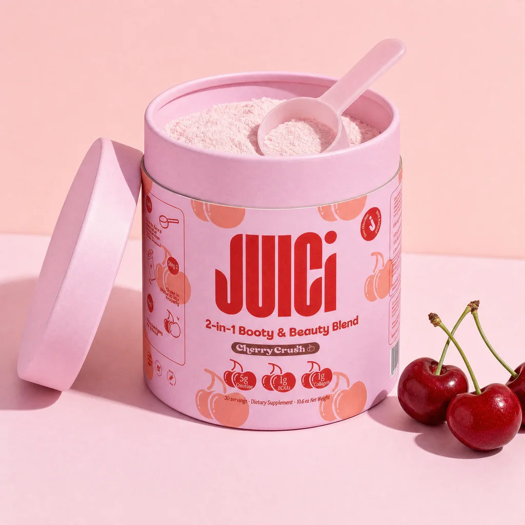 An open tub of JUICi Cherry Crush with its pink lid off, a pink scoop in the powder and fresh cherries in a pink studio